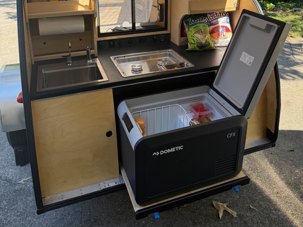 A Dometic CFX fridge on a slide pulled out of a teardrop galley bay, lid open and clear of the countertop, sink and cooktop above