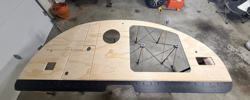 A CNC-cut teardrop side wall panel on sawhorses with a routed window opening, port hole, and wiring channels