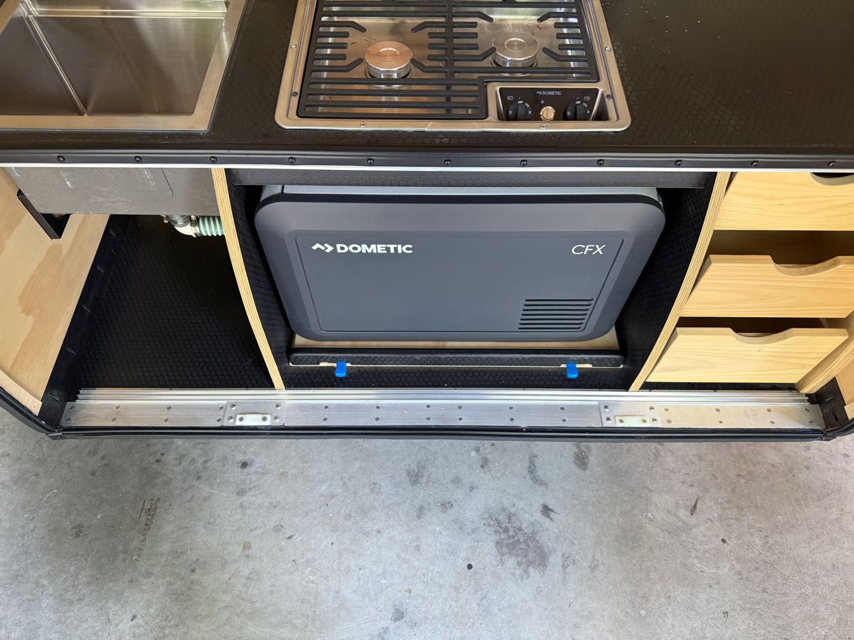Dometic CFX3 portable compressor fridge mounted on a slide in the lower galley bay of a teardrop camper, with the cooktop above it