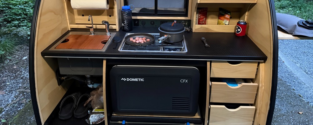A teardrop camper galley set up and cooking at a wooded campsite, with a Dometic CFX portable compressor fridge stocked in the bay below the counter