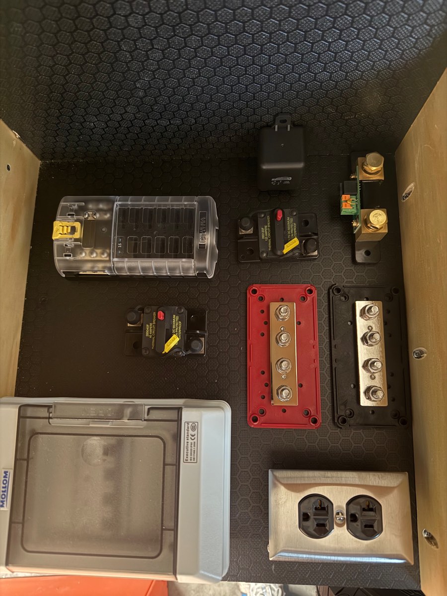 Electrical components staged and unmounted in the teardrop's power compartment — clear-cover blade fuse block, thermal circuit breakers, red and black heavy-duty bus bars, DIN-rail AC distribution box, shunt, and a duplex outlet