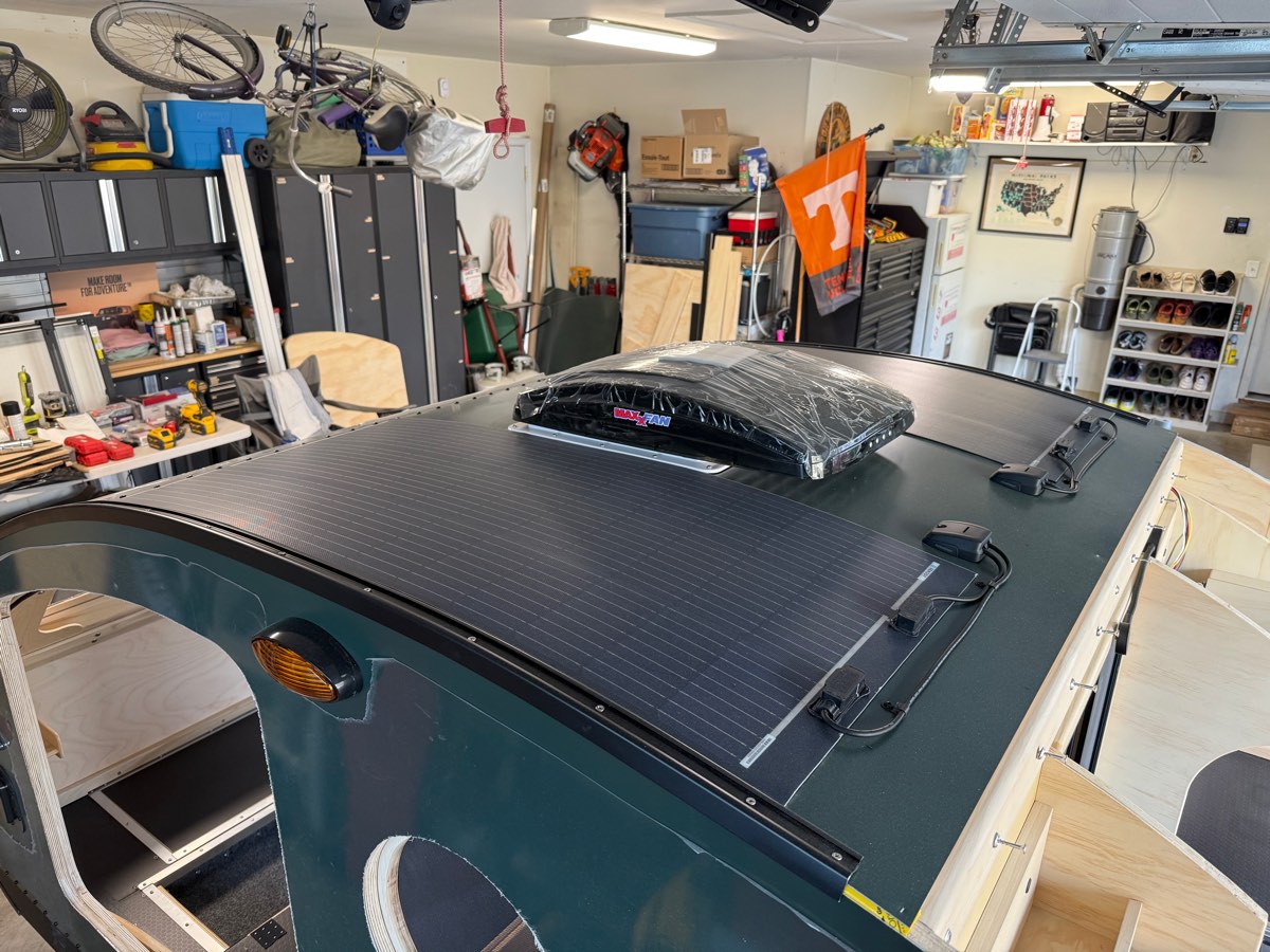 A flexible solar panel bonded to the curved roof of a teardrop camper with a powered vent fan ahead of it