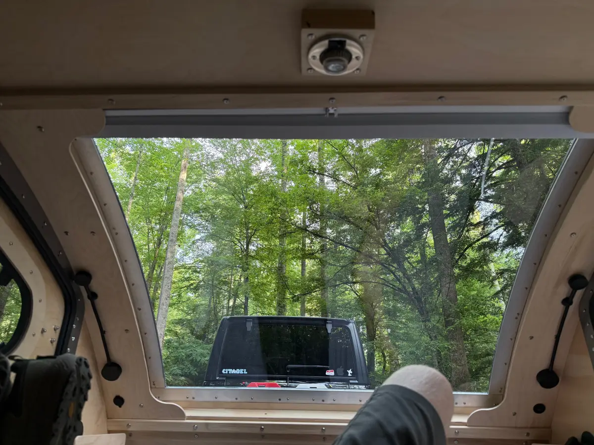 View from inside the teardrop camper looking out the open rear hatch window at the forest campsite, with feet propped up in the foreground