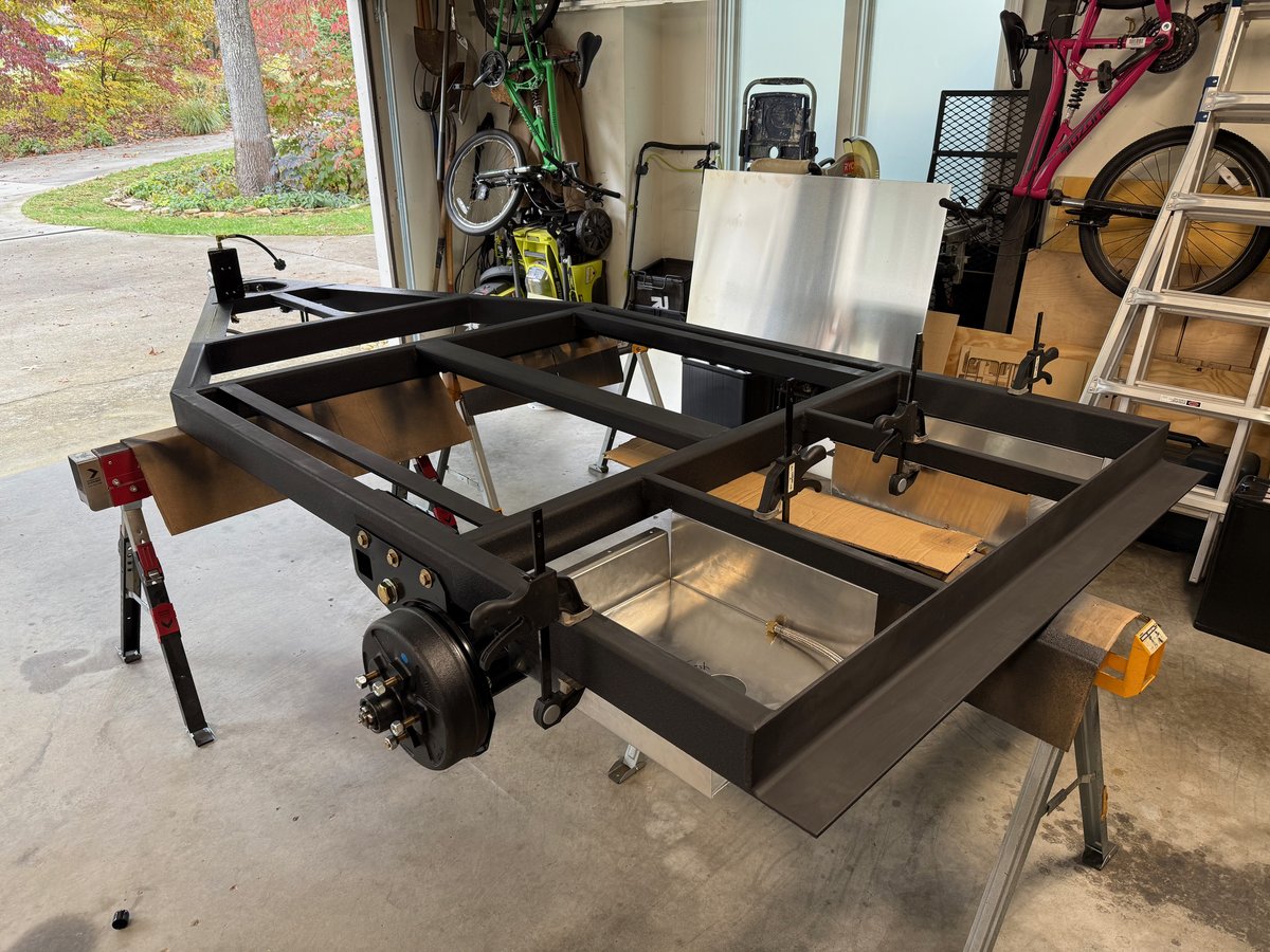 A Timbren axle-less unit bolted to the side of a black steel teardrop frame on sawhorses, a brake drum on its spindle and an aluminum tank pan behind it