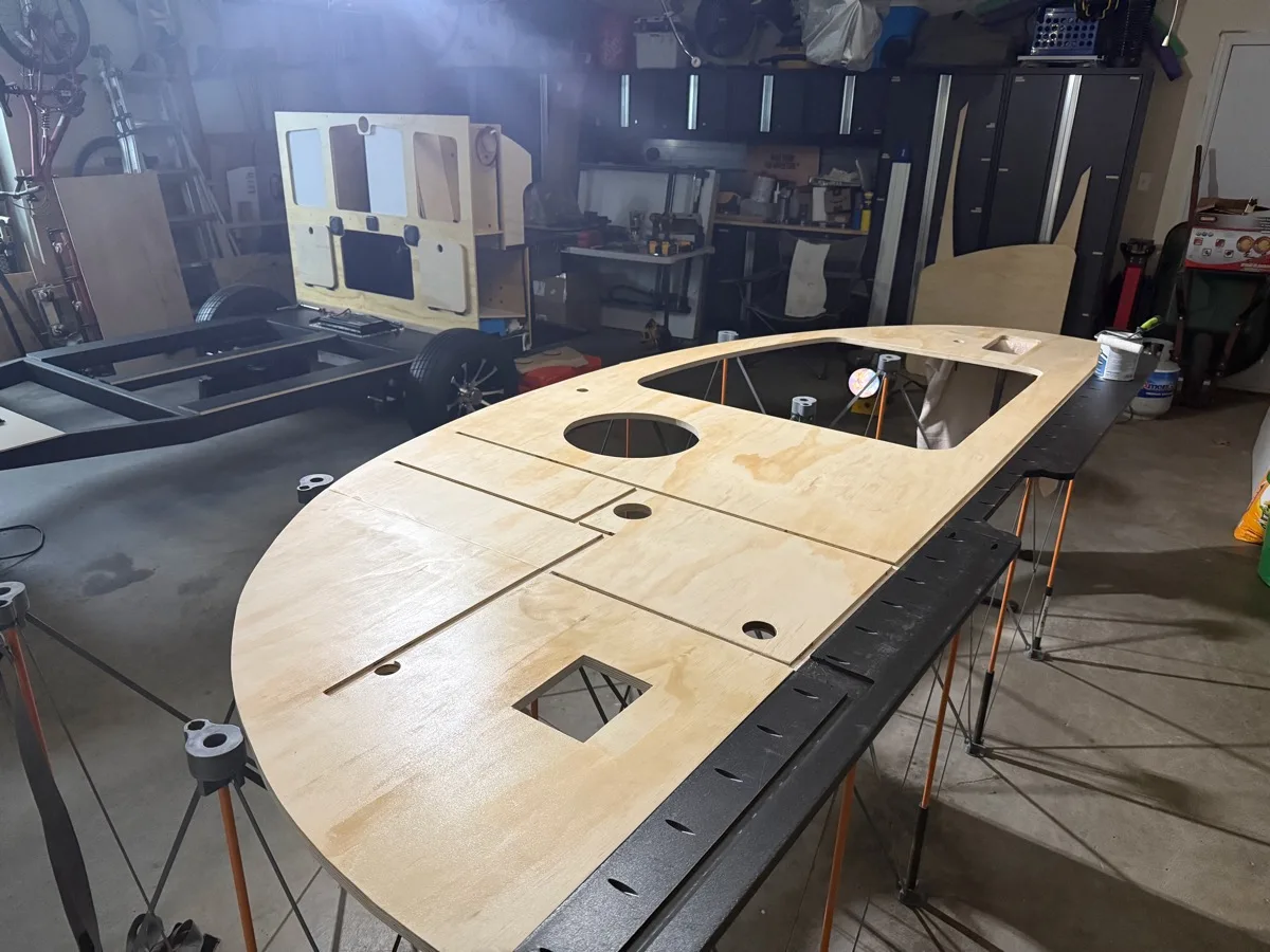 The interior side of a large curved plywood side wall panel with a window cutout and routed slots for the galley countertop and interior bulkhead, laid out on sawhorses in the shop
