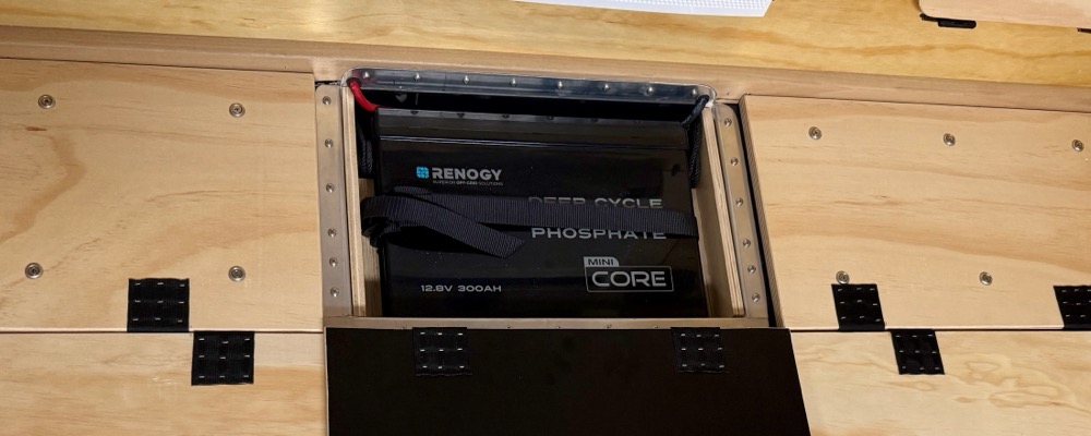 A Renogy Core Mini 12.8V 300Ah LiFePO4 battery strapped into a plywood-lined bay under the cabin floor, access hatch open