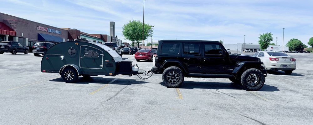 A black Jeep Wrangler hitched to a green teardrop camper in a parking lot, both sitting level