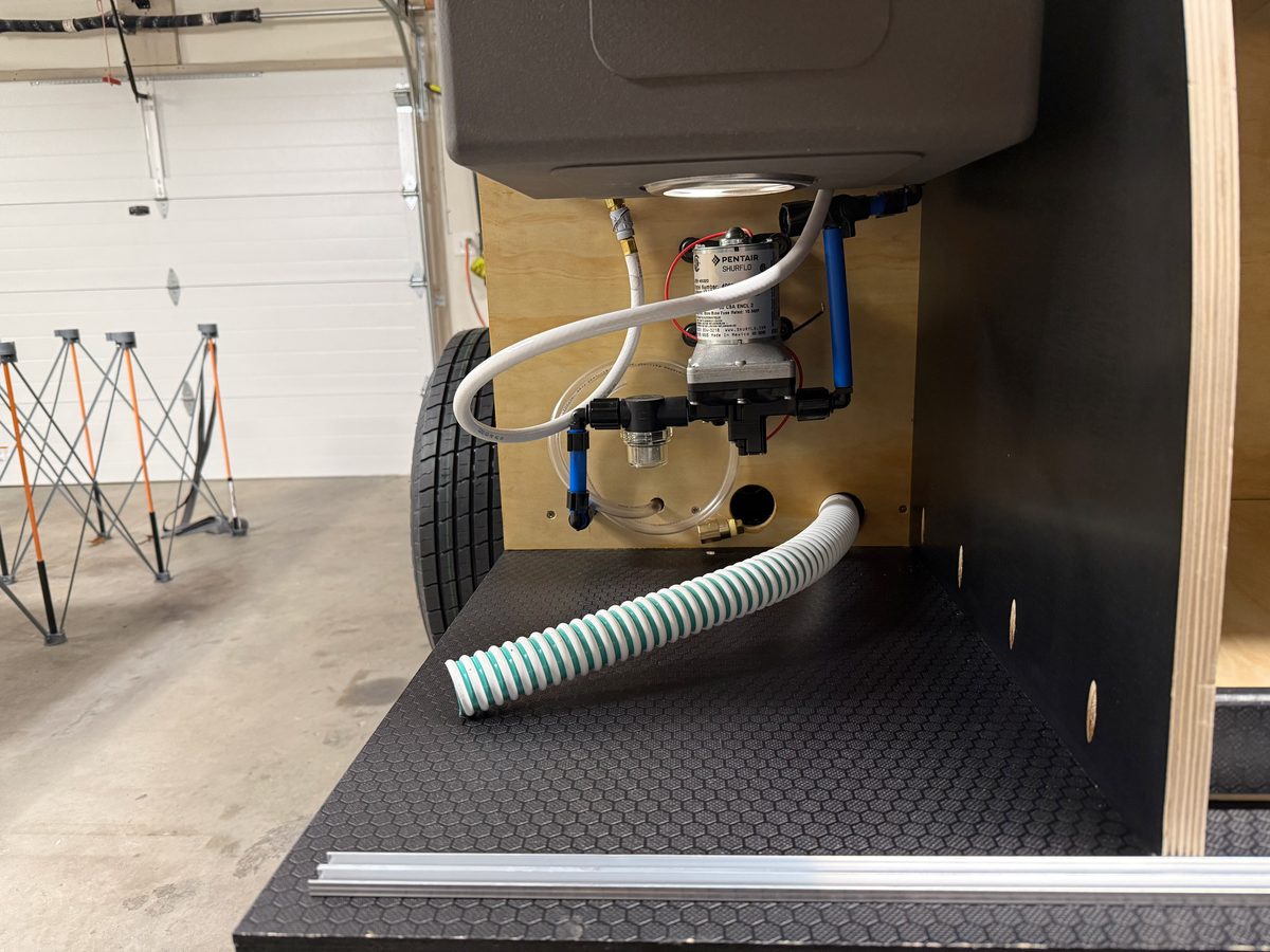 A Pentair SHURflo 12V water pump mounted on the plywood back wall under a teardrop galley sink, with a strainer, blue and white water lines, and a ribbed drain hose