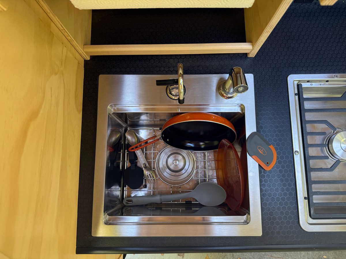 Looking down into a stainless teardrop galley sink with the stopper in the drain, a skillet, plates, a spork and a spoon on a wire rack, the faucet and sprayer behind