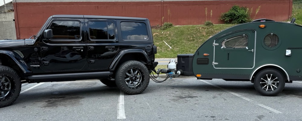 A green teardrop camper hitched behind a black Jeep Wrangler, showing the tow stance and wheel track of both