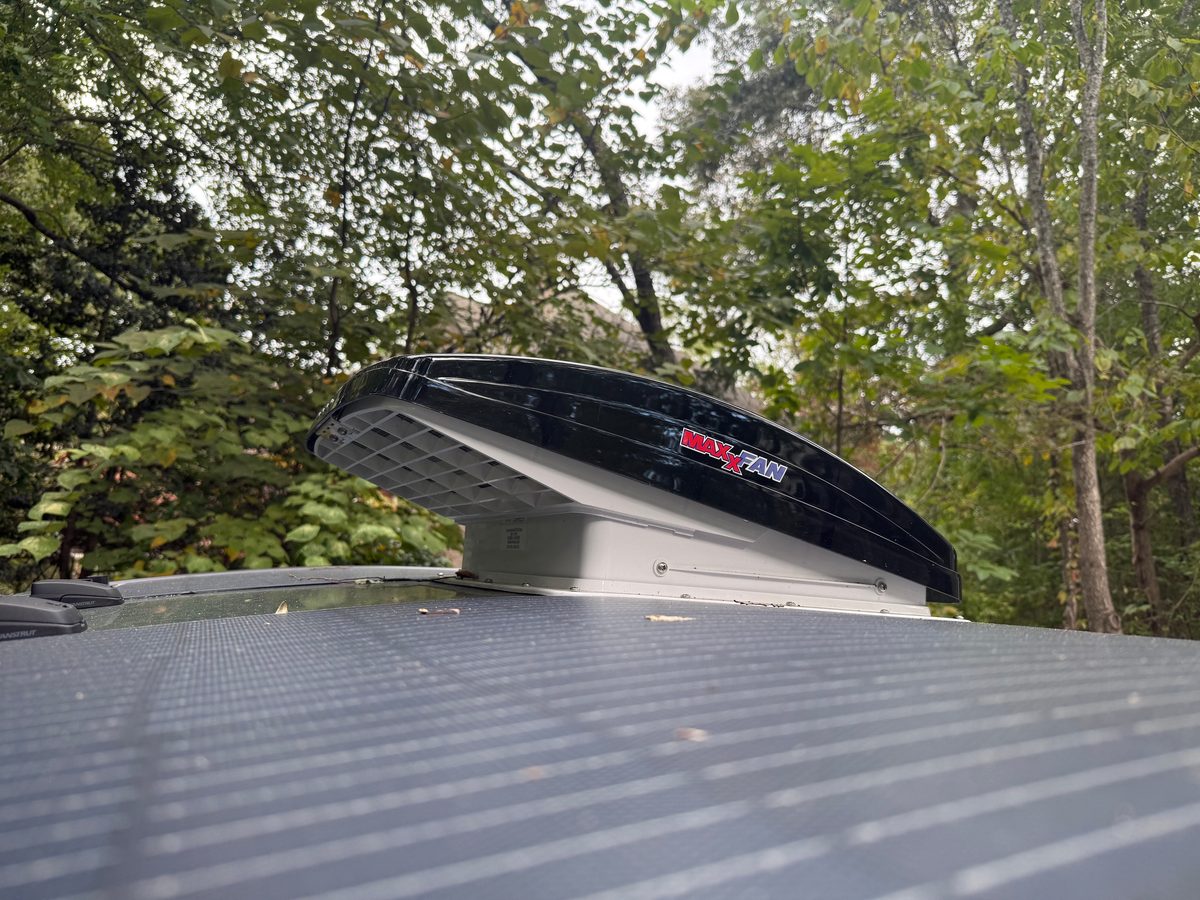 A black MaxxFan lid propped partly open on the roof of a teardrop, next to a flexible solar panel, with trees behind