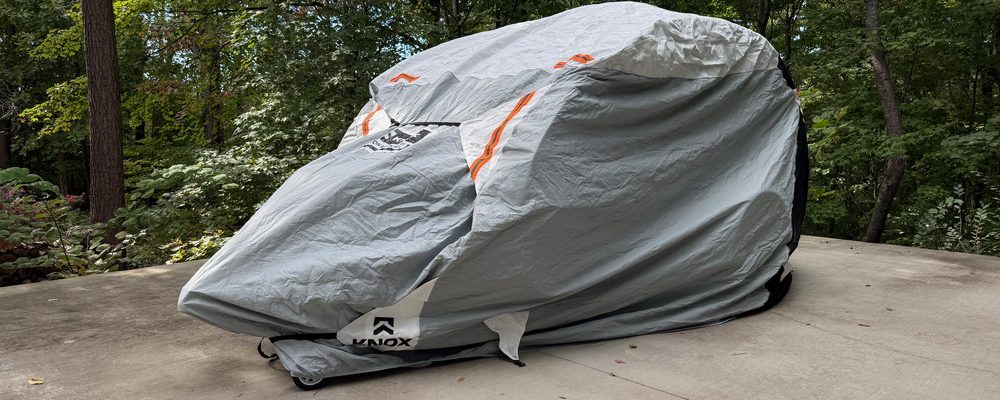 A teardrop trailer under a fitted gray cover on a concrete pad, with the woods behind it